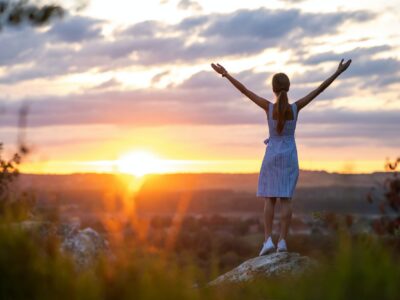 Dark silhouette of a young woman standing with raised up hands on a stone enjoying sunset view