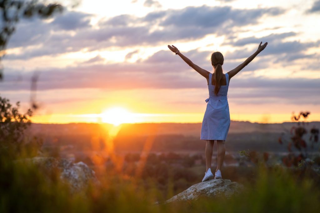 Dark silhouette of a young woman standing with raised up hands on a stone enjoying sunset view