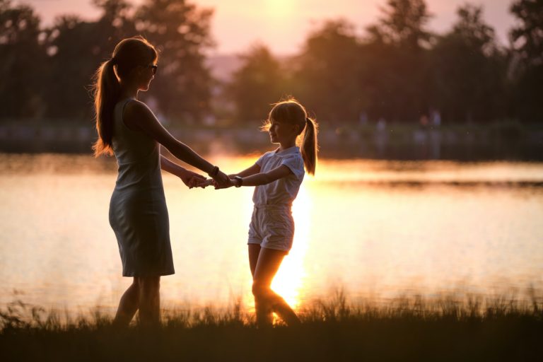Happy mom and daughter girl relaxing holding hands enjoying time together in summer park at sunset